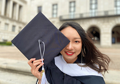 graduating student in gown holding mortarboard over eye